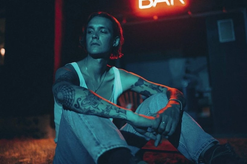 Tattooed man sitting outside a bar at night under the red neon sign, wearing a white tank top and blue jeans, looking off camera.