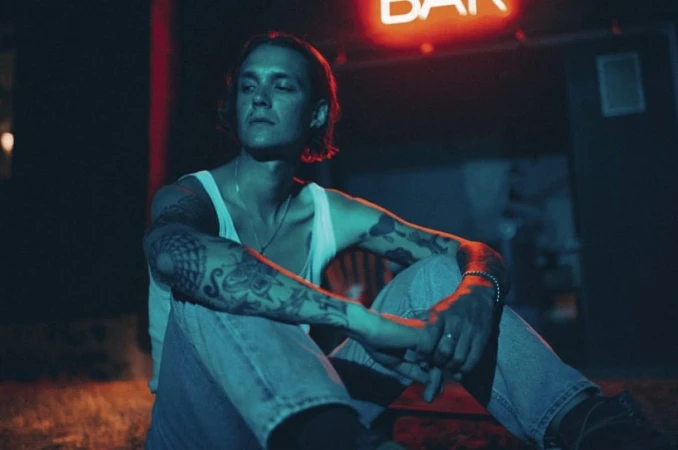 Tattooed man sitting outside a bar at night under the red neon sign, wearing a white tank top and blue jeans, looking off camera.