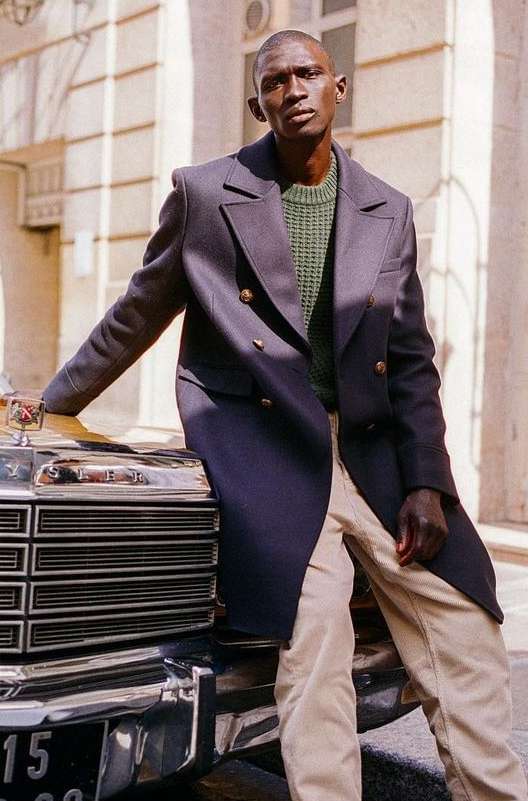 Dark-skinned male model poses next to a vintage car in a navy overcoat, light pants, and green sweater, set against an urban backdrop.