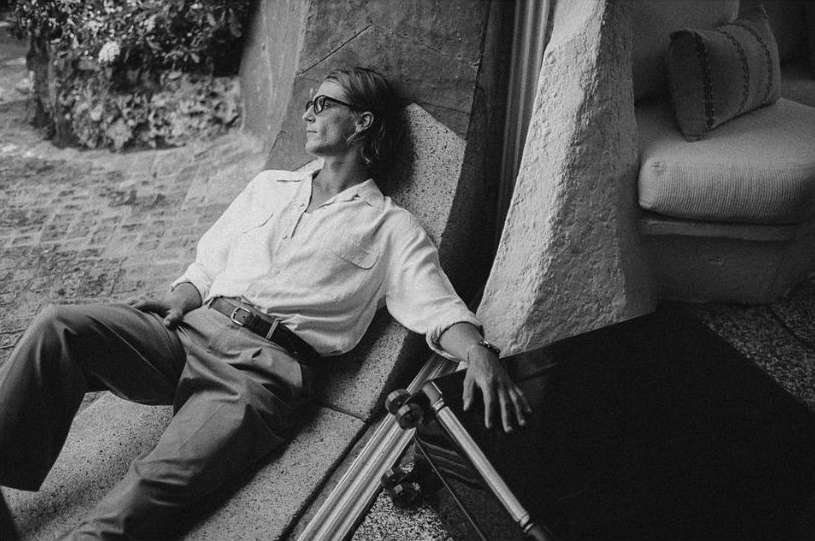 Adult male with glasses in a white shirt and grey pants leaning against stone wall with carved details near neutral colored furniture.