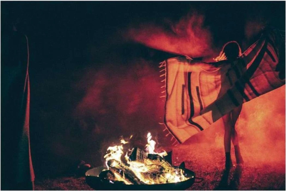 Person in silhouette standing near a campfire, holding blanket with black stripes, bathed in a red and orange glow at nighttime.