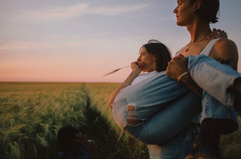 Man carrying woman in field at sunset. Couple in embrace in rural setting with tall grasses and pink and yellow sky. Romance and nature.