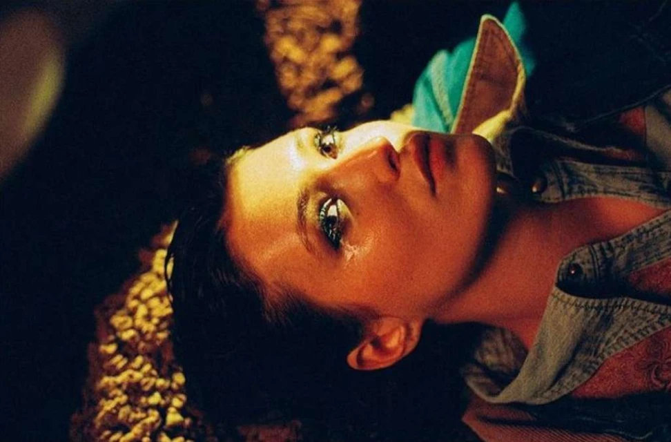 Young woman lying down looking up with a tear on her cheek. She is wearing a denim jacket with a plaid shirt underneath.