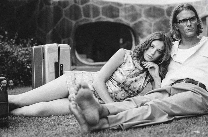 A young couple relaxes on the grass with a vintage suitcase in this black and white outdoor shot. The woman reclines near her partner.