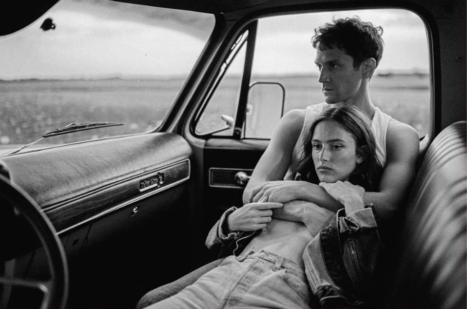 Monochrome portrait of a couple in an older pickup truck, the man embraces the woman, open field visible through the windshield.