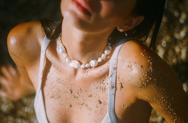 Woman with pearls lying on sandy beach, showcasing an elegant pearl necklace and matching earring in sunlit, natural setting.