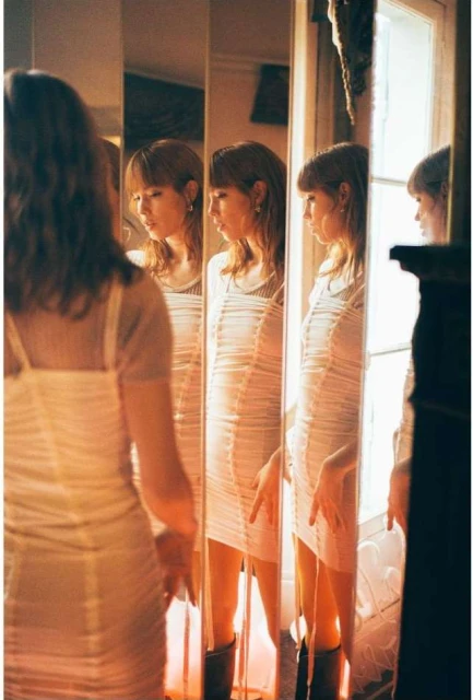 Young woman gazes into a tall mirror, reflecting her multiple times; she wears a white dress and black boots in a warmly lit interior.