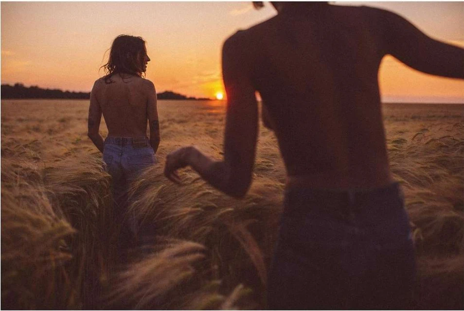 Two women are standing in a field of golden wheat at sunset. The woman in the foreground is closer to the camera.