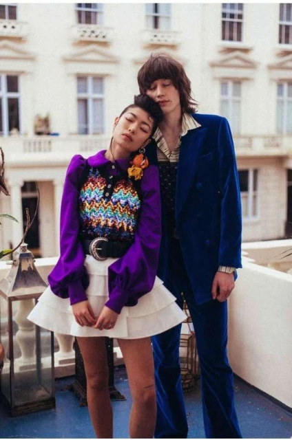 Two people pose in front of a building. An adult female wears a purple top and white skirt, and an adult male wears a blue suit.