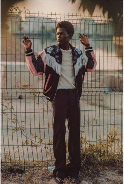 Adult male model posing in fashionable Western-style jacket against a wire fence backdrop in an outdoor environment with natural light.