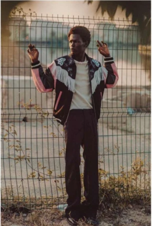 Adult male model posing in fashionable Western-style jacket against a wire fence backdrop in an outdoor environment with natural light.