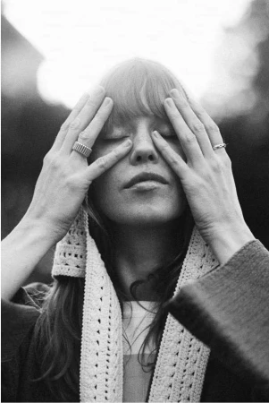 Black and white portrait of a woman with bangs holding her hands to her face covering her eyes, wearing a knitted scarf.