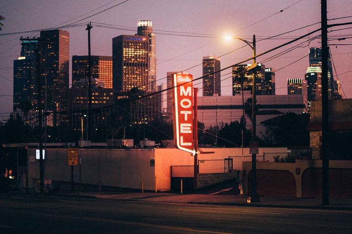 Illuminated 'Motel' sign glows against a city skyline at dusk, power lines cross the sky, and a dark street in the foreground.
