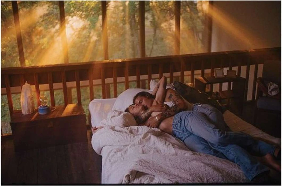 A couple is lying in bed near a window with natural light streaming through, creating a warm and intimate atmosphere in the bedroom.