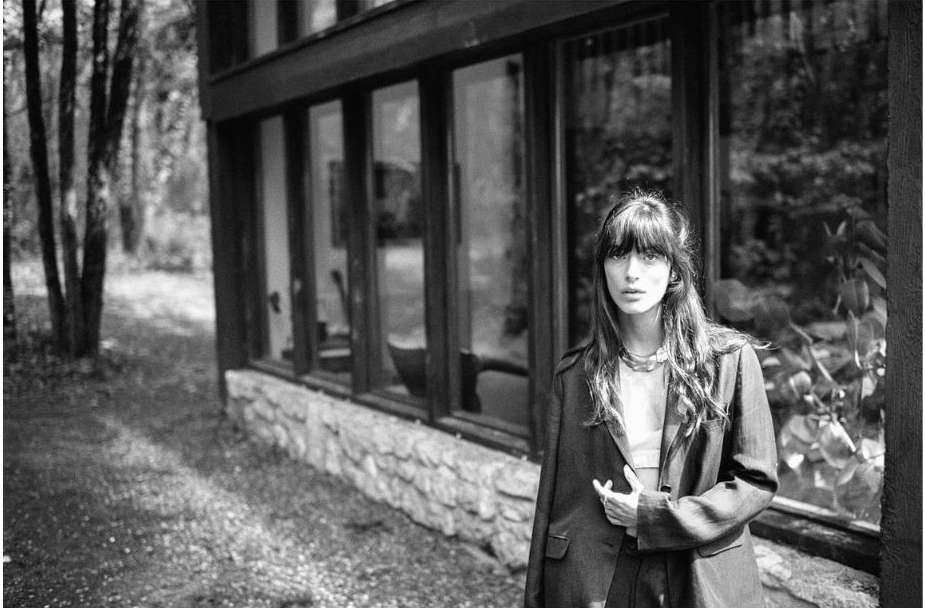 A black and white medium shot of a woman in a blazer standing outside a stone building with large windows and a forest in the background.