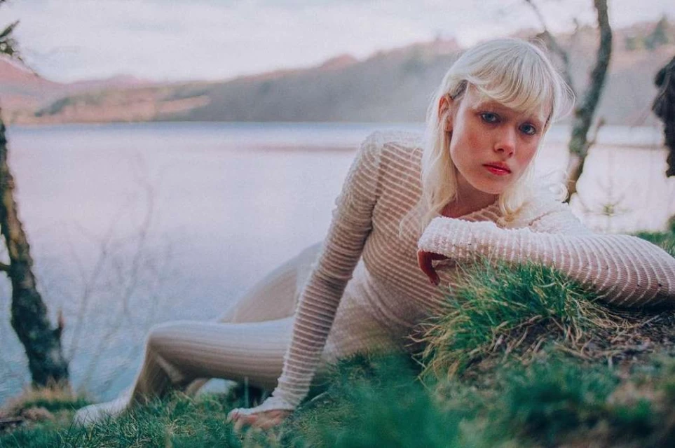 Woman with platinum blonde hair poses by lake in natural light wearing a textured long-sleeve top and pants; trees and mountains in distance.