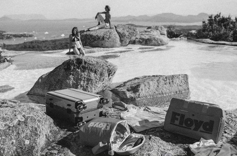 Two women relaxing near luggage by still water with a coastal view; one sits on rocks, the other kneels, black and white, travel theme.