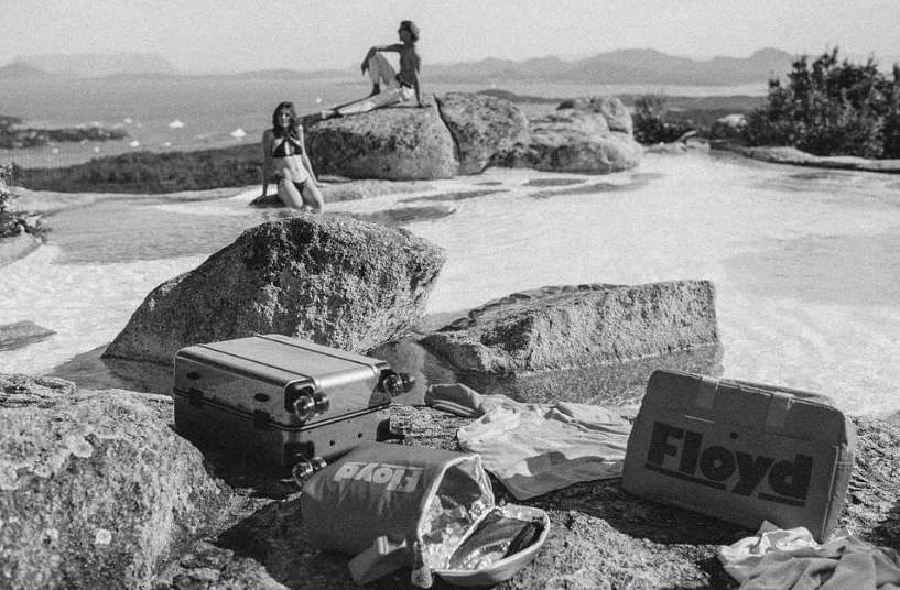 Two women relaxing near luggage by still water with a coastal view; one sits on rocks, the other kneels, black and white, travel theme.