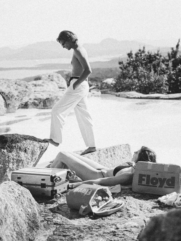 Monochrome shot of a shirtless man in white pants walking near a woman sunbathing with suitcases in a scenic outdoor landscape.