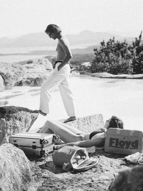 Monochrome shot of a shirtless man in white pants walking near a woman sunbathing with suitcases in a scenic outdoor landscape.