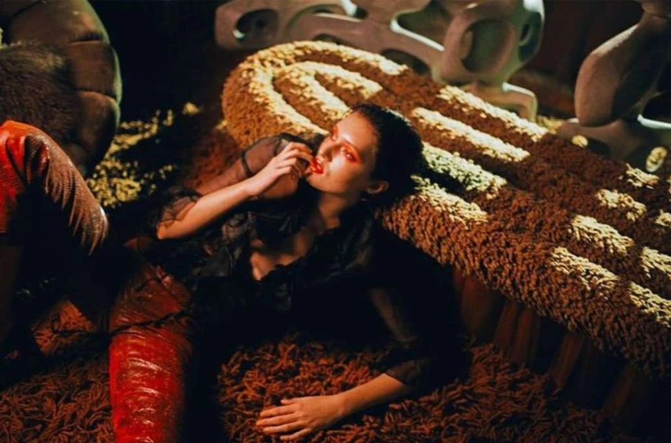 A woman in dark clothing lies on a shag rug with red lips, pale skin, and red eye shadow with sculptural forms in the background.