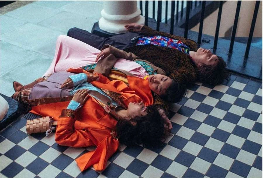 Three young adults casually lounge on a checkered floor in stylish attire near a white column, creating a fashionable and relaxed scene.