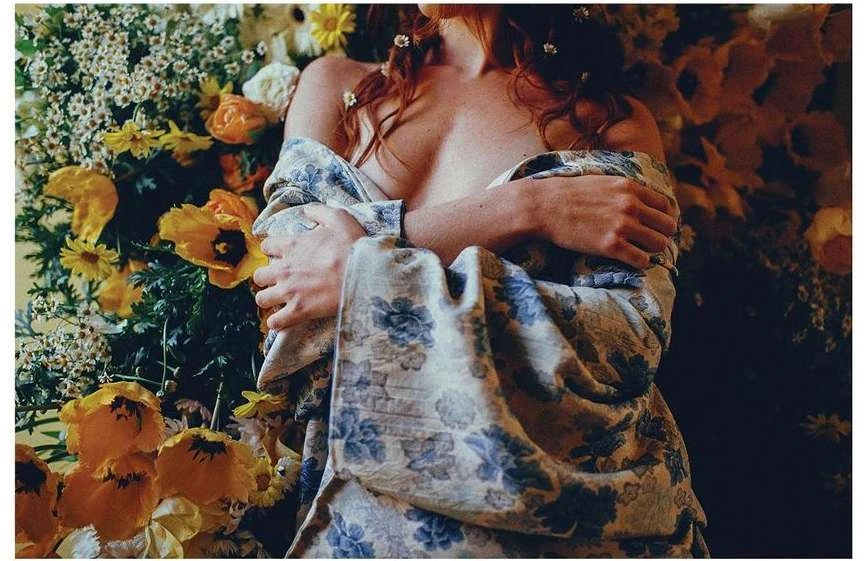 Red-haired woman lies among vibrant orange and white flowers wearing a floral-patterned shawl, creating a sensual floral portrait.