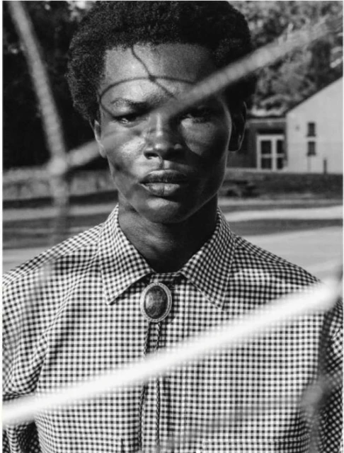Black and white portrait of an adult with a patterned shirt and bolo tie, looking directly at the camera through a blurred foreground.