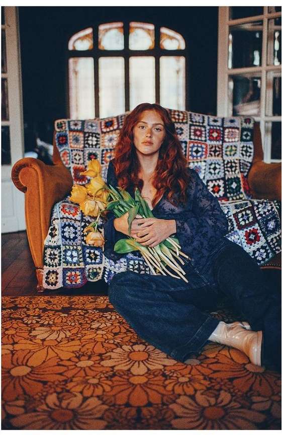 Young woman with red hair sits on a floral rug with yellow tulip bouquet in front of a couch with a vintage quilt.