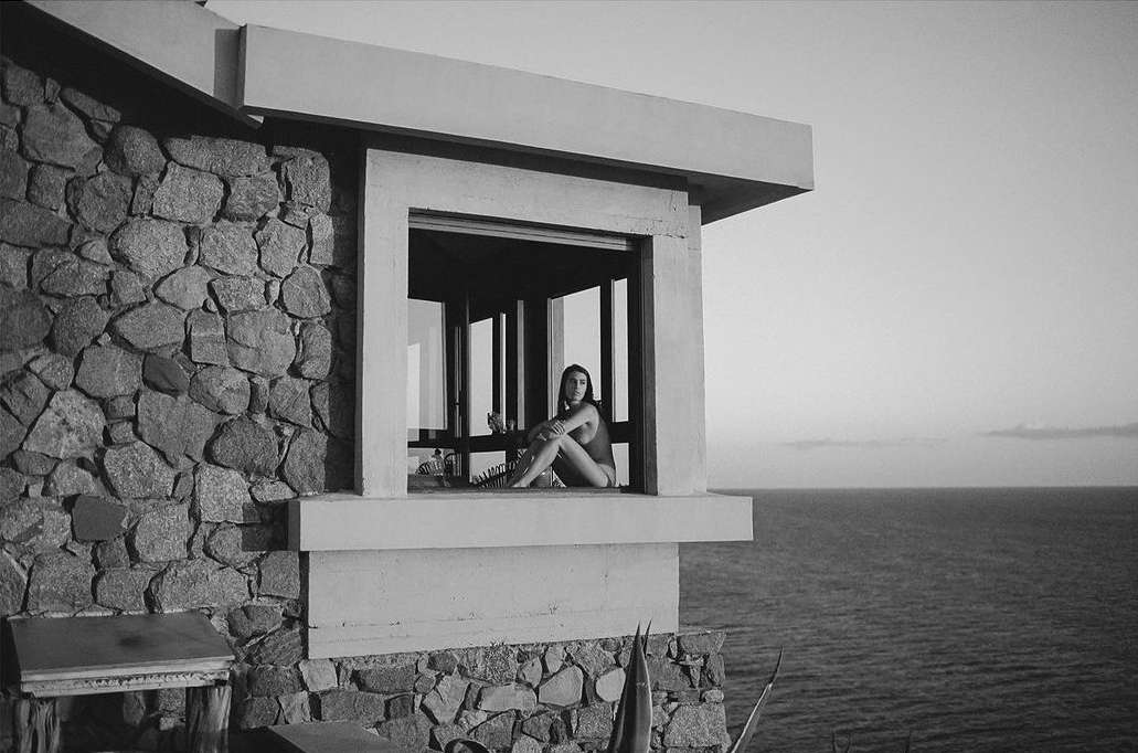 Woman in swimsuit sits in window of stone building overlooking ocean. Monochrome image. Building's stone texture contrasts with smooth water.
