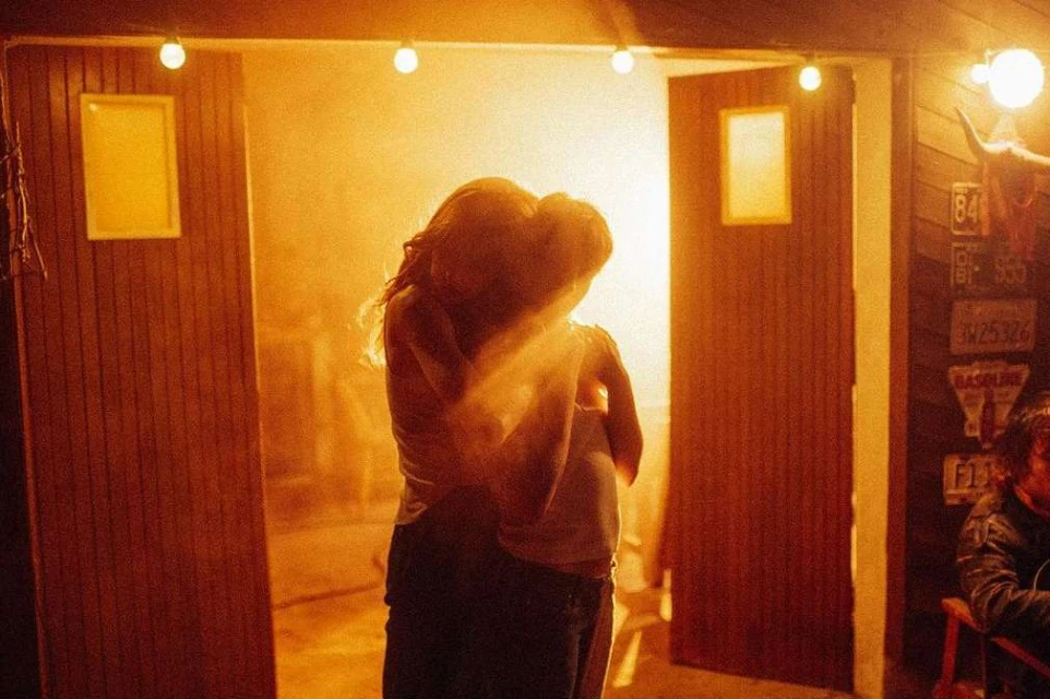 A warmly-lit scene of a couple embracing in a doorway. Interior lights and wall details are subtly visible in a vintage style.