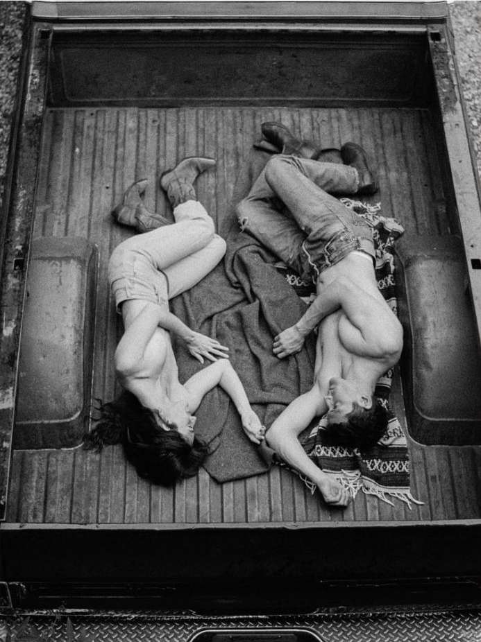 Black and white aerial shot of two shirtless humans, a male and female, lying in the back of a truck holding hands on a blanket.
