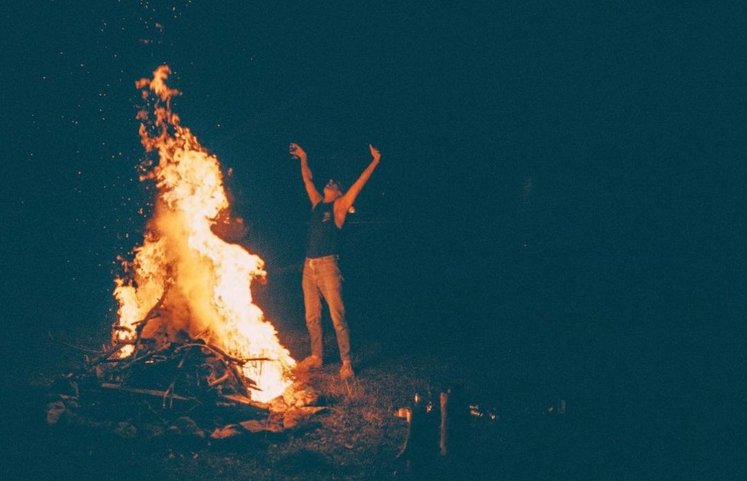 Person standing with arms raised near a large campfire at night, with dark sky and sparse vegetation.