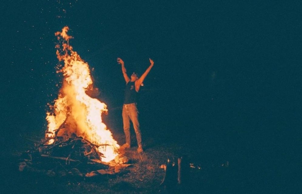Person standing with arms raised near a large campfire at night, with dark sky and sparse vegetation.