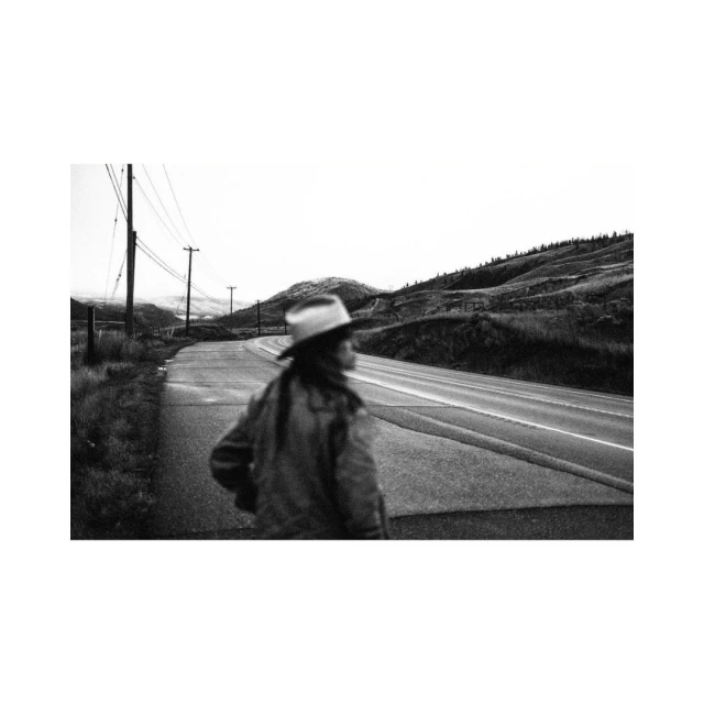 Monochrome image of a blurred female figure in a hat walking on a road, with hills and power lines visible in the background.