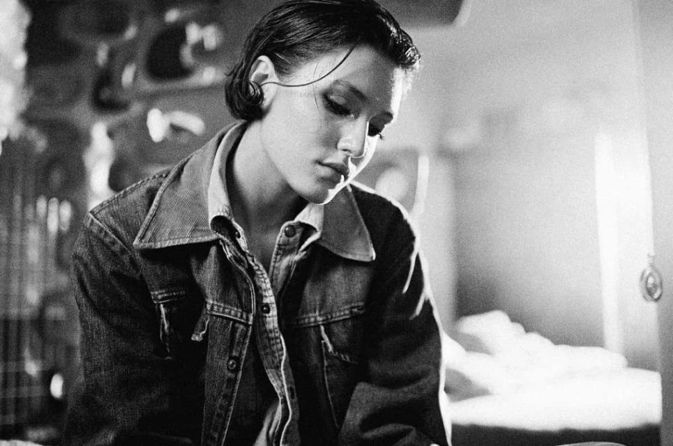Black-and-white portrait of a young woman in a denim jacket, head tilted, indoors with natural light and a soft, classic aesthetic.