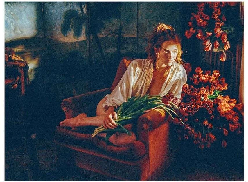 Woman in a white shirt sits in a red armchair, holding a bouquet of tulips, with a painted backdrop creating a moody, artistic portrait.