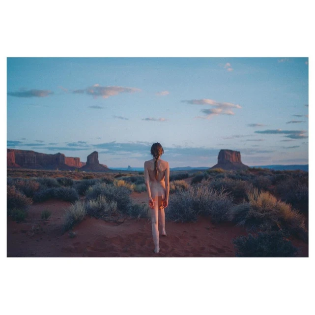 Nude woman with braided hair walks in the desert toward monumental sandstone buttes beneath a blue sky at dusk in a landscape photograph.