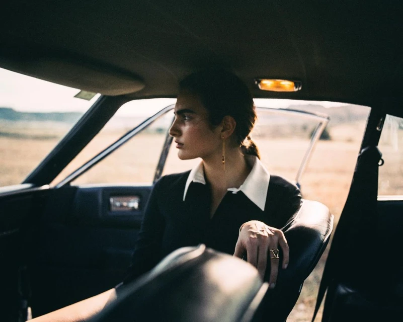Adult female sits inside classic car with desert landscape visible through open door, wearing black and white collared dress and jewelry.
