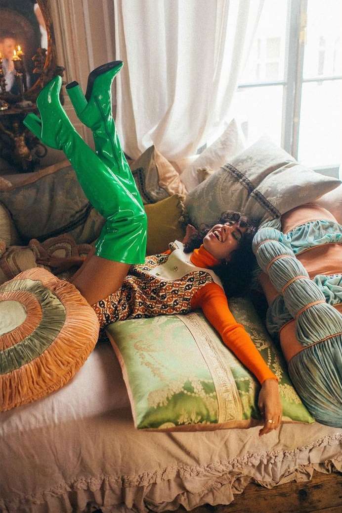 Smiling woman in orange top and patterned dress reclines among decorative pillows wearing bright green boots by a window.