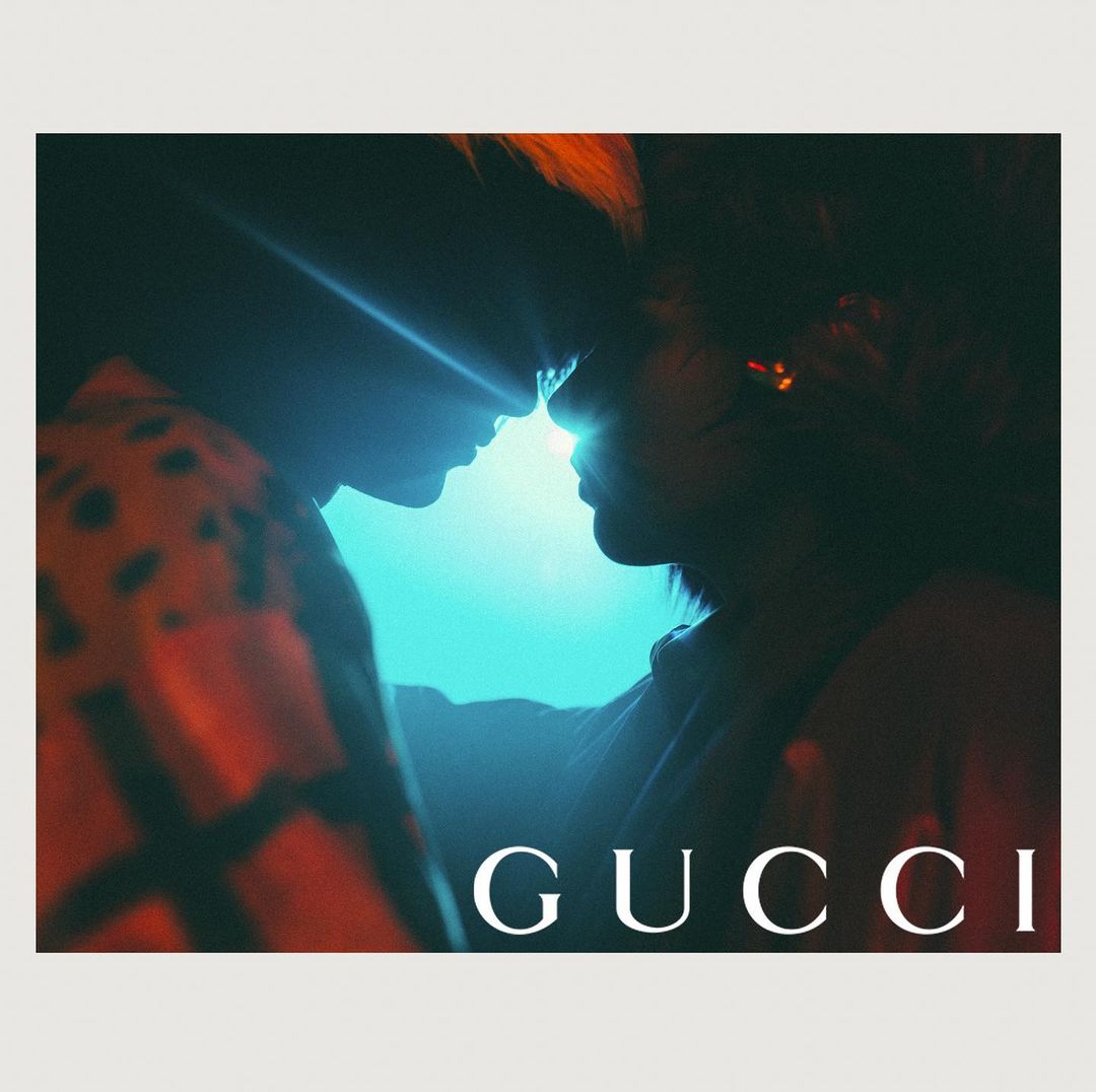Two people facing each other closely under dramatic blue and red lighting with the word GUCCI visible below