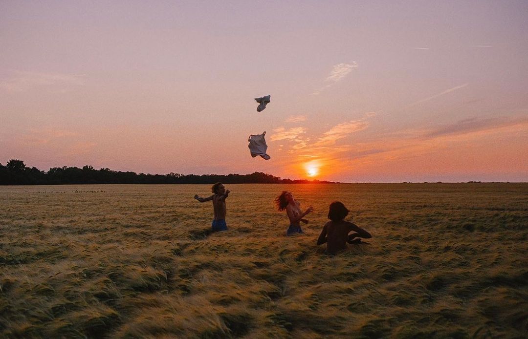 Four shirtless children stand in a golden wheat field at sunset, with clothing items tossed in the air near the horizon.
