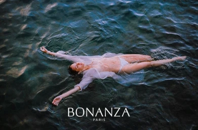 Woman floating on her back in dark water wearing a white sheer shirt and white bikini bottoms by photographer Theo Gosselin