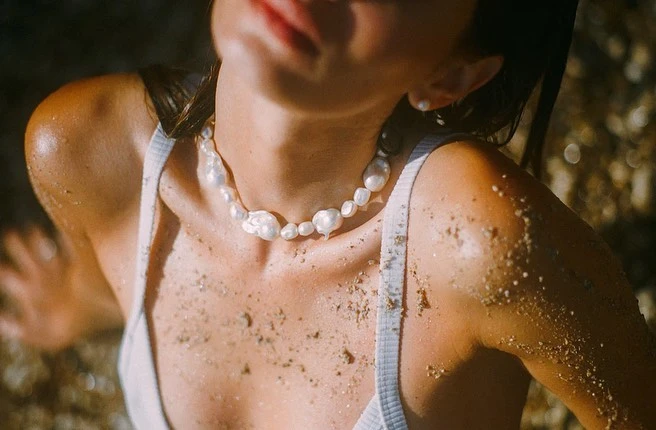 Close-up portrait of a woman wearing a white ribbed tank top and a baroque pearl necklace, with sand dusted across her skin outdoors.