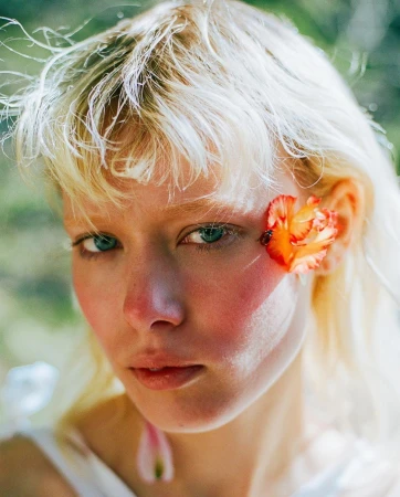 Close-up portrait of a blonde woman with blue eyes wearing an orange flower tucked behind her ear, photographed by Theo Gosselin.