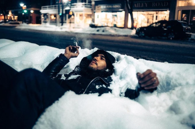 Man in black jacket lying in snow at night holding a cigarette with street lights glowing in background