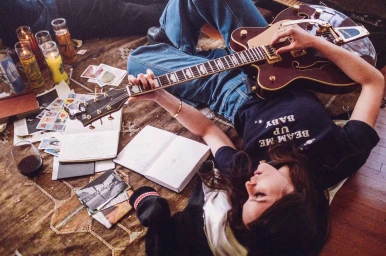 A woman lying on a wooden floor playing an electric guitar, surrounded by open notebooks, glass candles, and scattered papers.