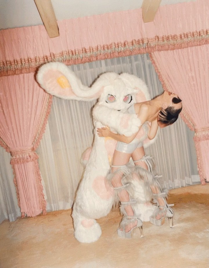 Woman in shiny outfit hugs a large, white furry bunny costume against a frilly, light pink background in an eye-level, medium shot.