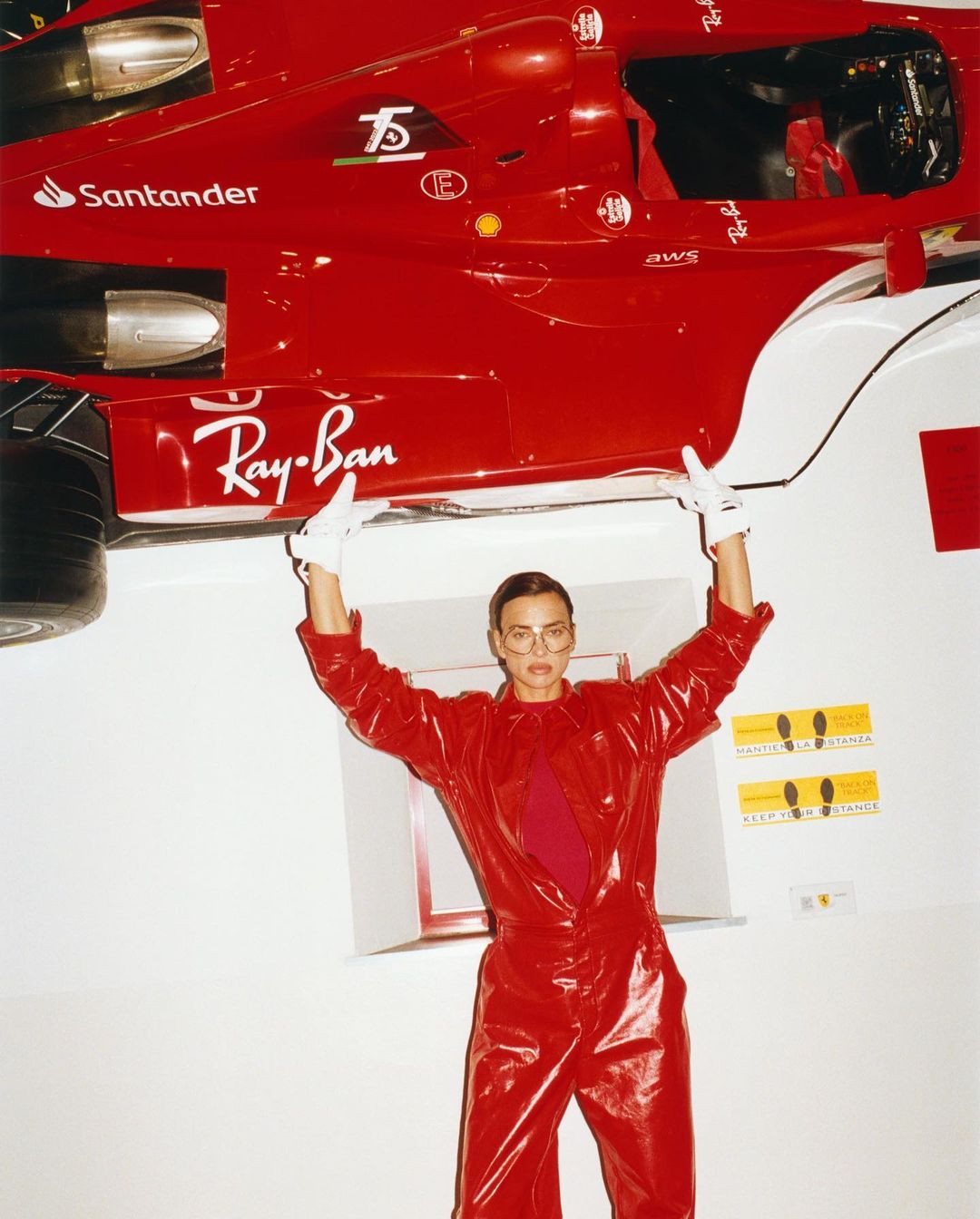 Female in a glossy red jumpsuit holds up a Formula 1 race car in a bright studio setting, showcasing Ray-Ban and Santander logos.