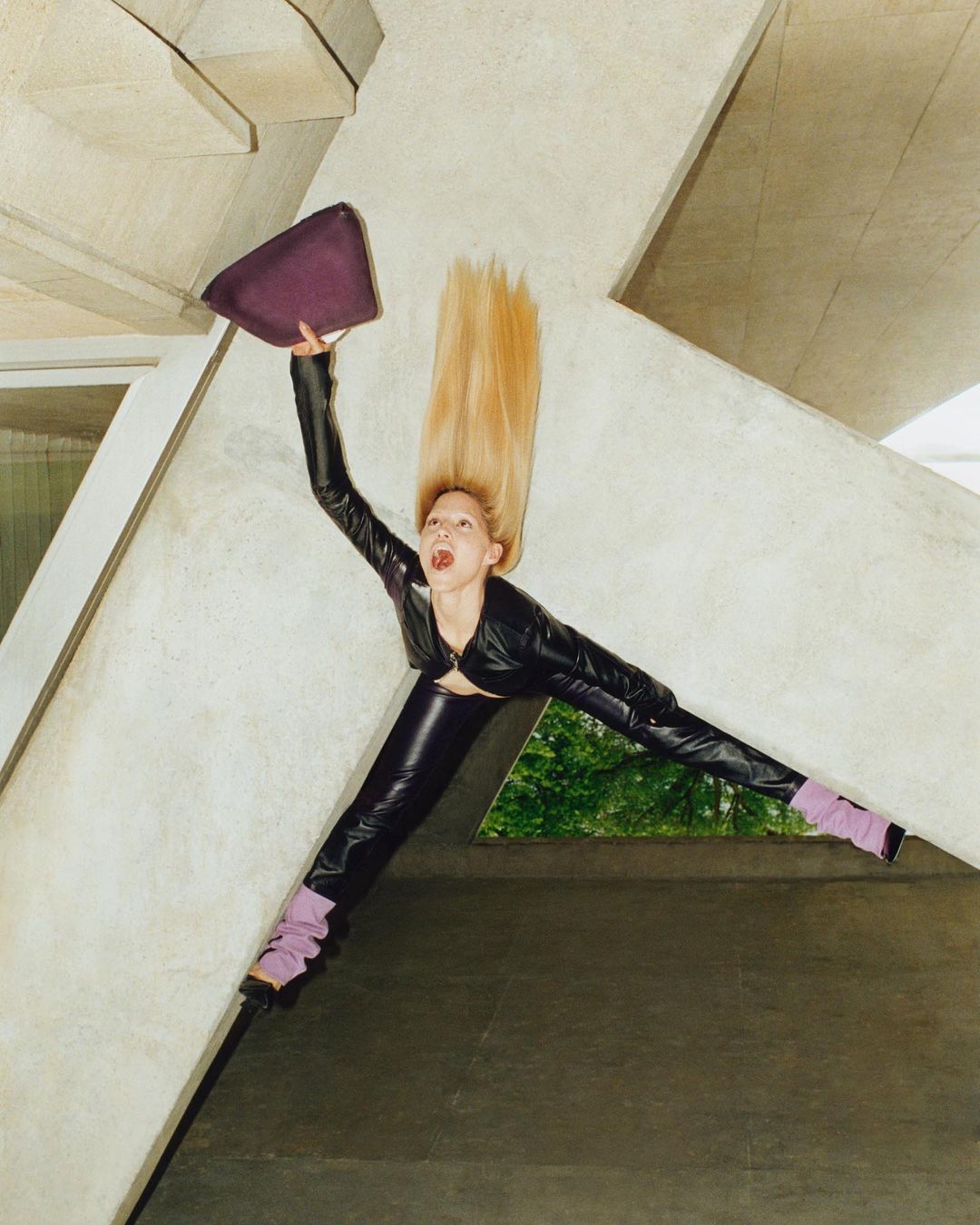 Woman in black pleather outfit raises purple purse in studio with geometric concrete background; long blonde hair, fashion shot.